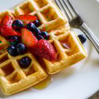 Golden brown waffles steam on a plate, topped with fresh berries and drizzled with warm maple syrup for a classic American breakfast.