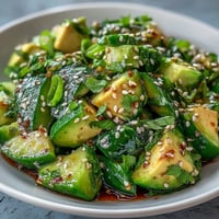 A vibrant bowl of smashed cucumber and avocado salad with sesame seeds and green onions, dressed in a tangy sesame-ginger vinaigrette.